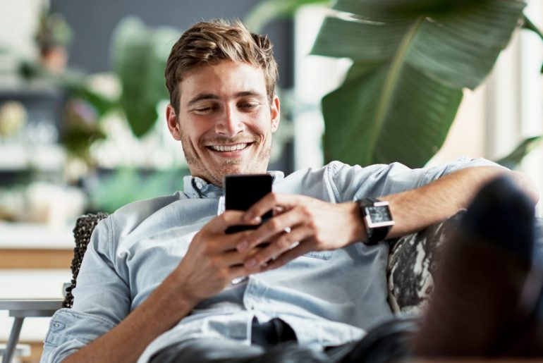 Mann liegt lächelnd auf der Couch mit seinem Handy in der Hand